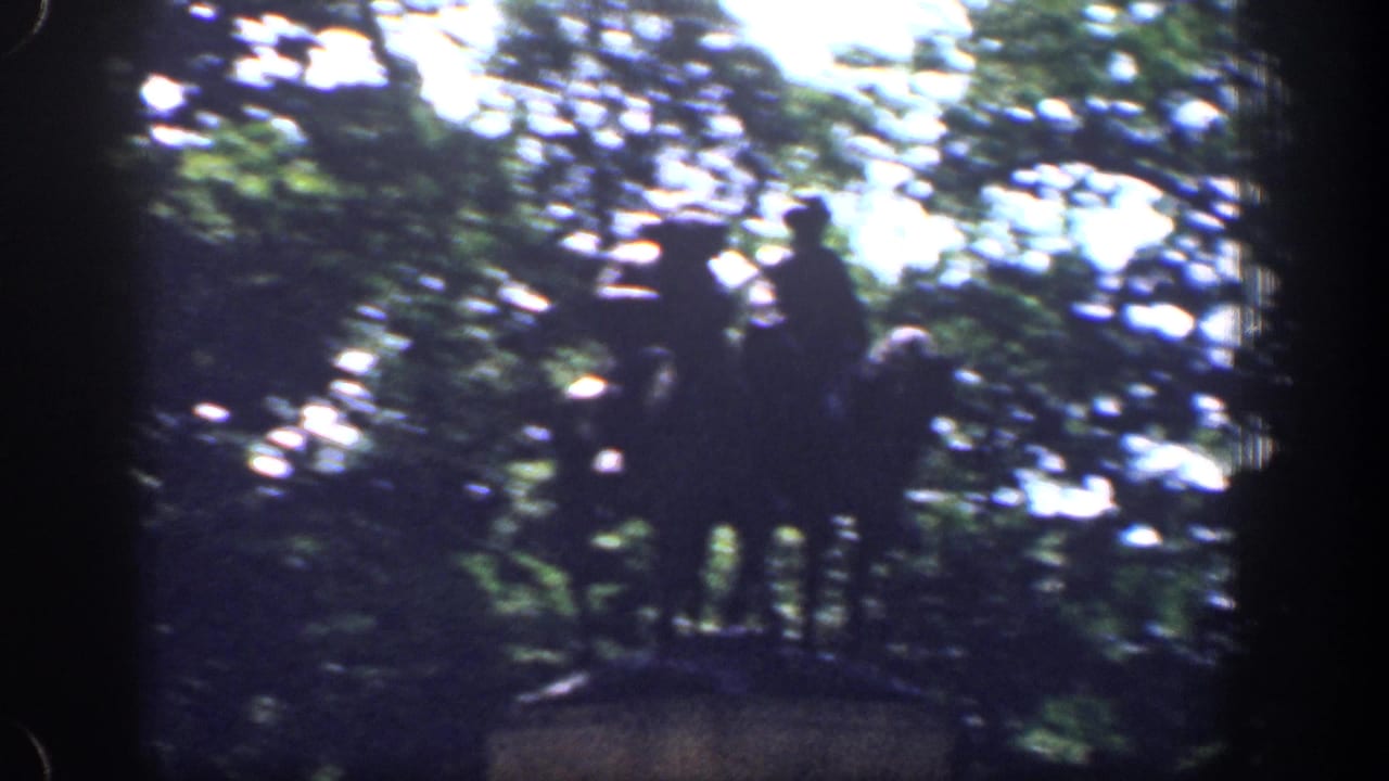 The drive-by camera passes a dark mounted monument, with the figures silhouetted against trees and bright sky.