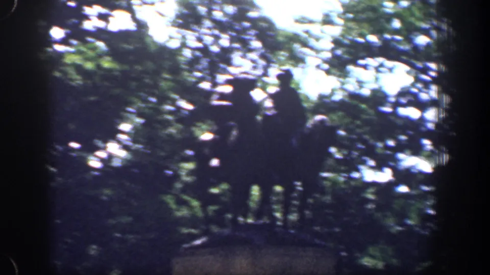 The drive-by camera passes a dark mounted monument, with the figures silhouetted against trees and bright sky.