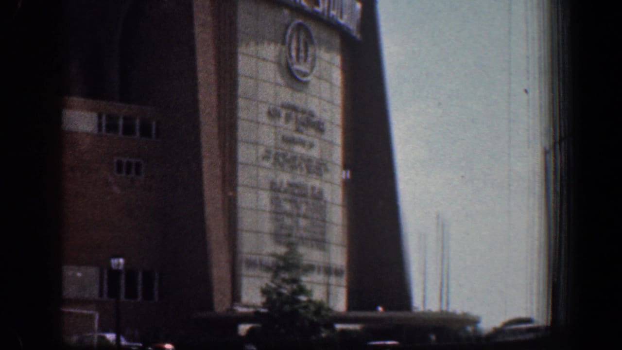 1961 Washington, DC home-movie frame showing Memorial Stadium entrance signage, brick walls, a small tree, and parked cars below.