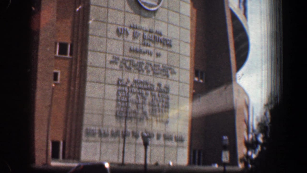 Tall building facade with inscription, brick sides, curved upper structure, and street trees.