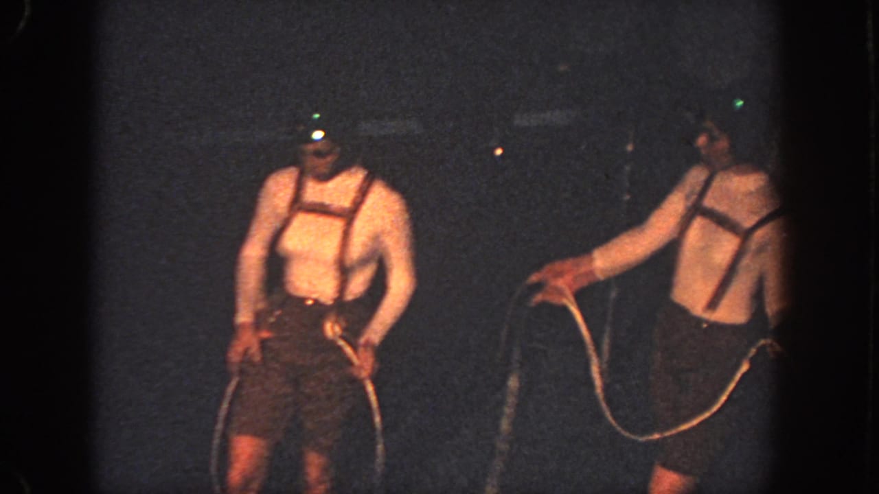 Two costumed swimmers in a dark pool hold ropes while wearing lighted headgear.