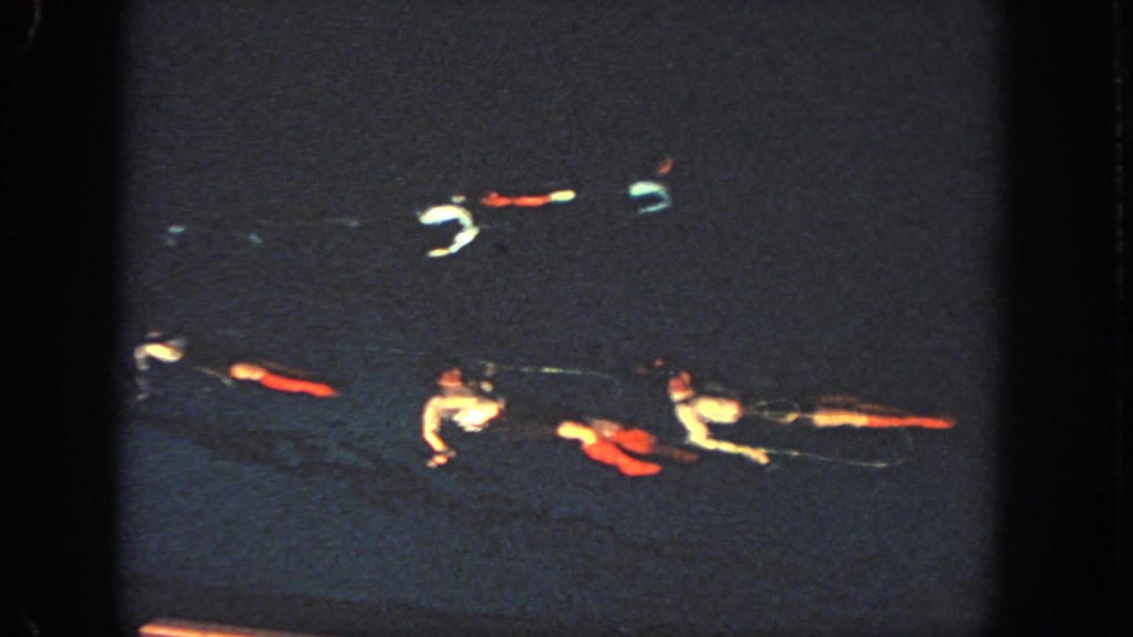 1951 Georgia home-movie frame showing swimmers lying or floating in formation, with pale shirts and colored costume pieces visible against dark pool water.