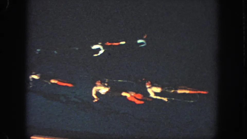 1951 Georgia home-movie frame showing swimmers lying or floating in formation, with pale shirts and colored costume pieces visible against dark pool water.
