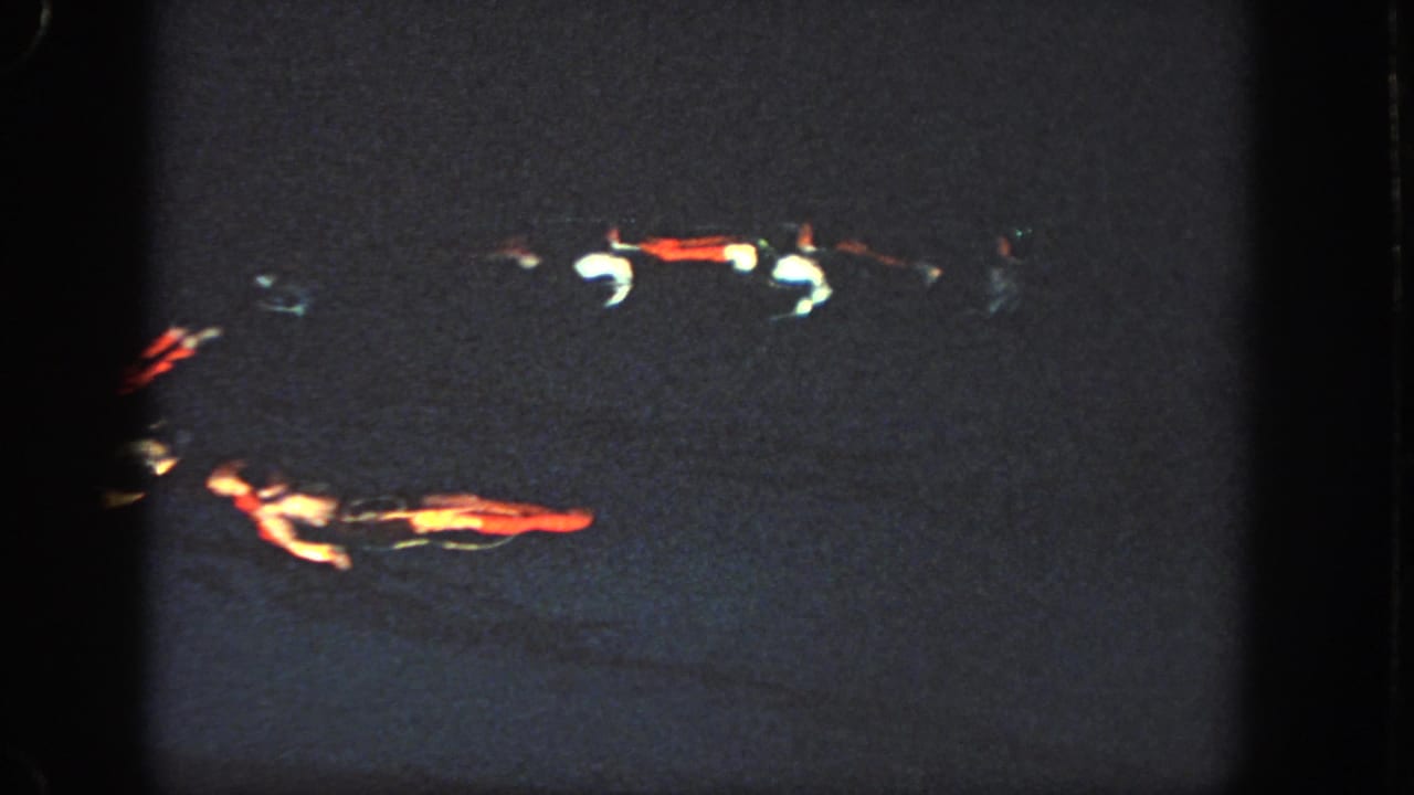 A low-light 8mm frame shows multiple swimmers spaced across the dark pool, with illuminated red and white costume details visible.