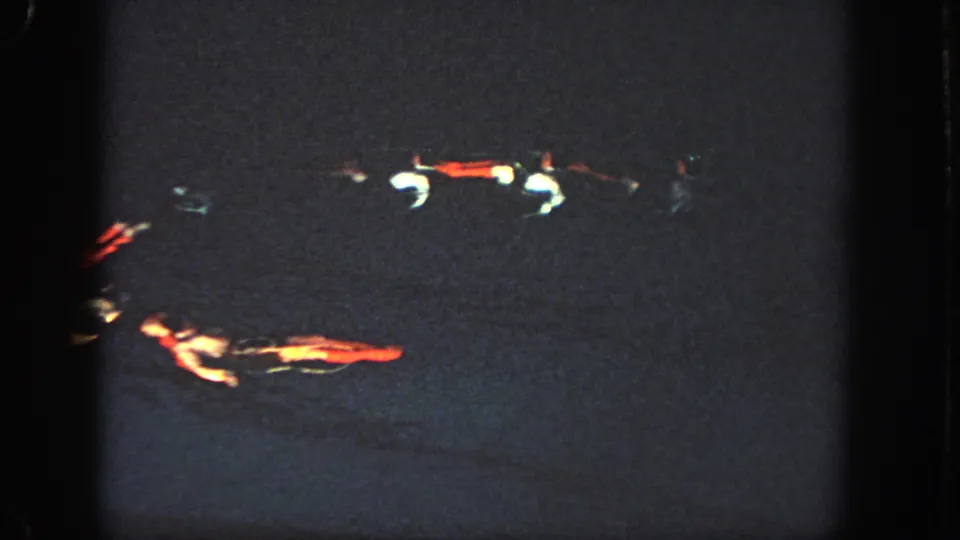 A low-light 8mm frame shows multiple swimmers spaced across the dark pool, with illuminated red and white costume details visible.