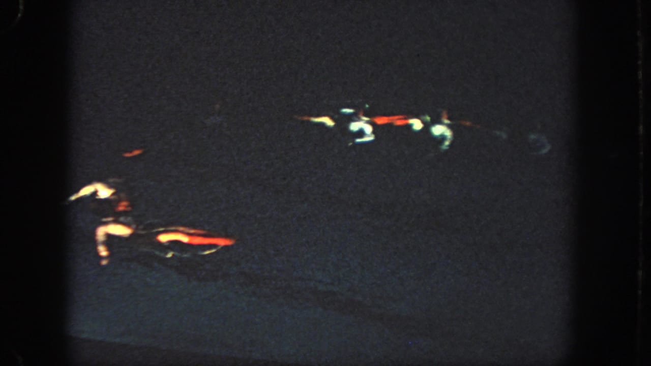 Near the end of the clip, two swimmers are partly illuminated in the dark pool, with red and white costume details standing out in the 8mm frame.