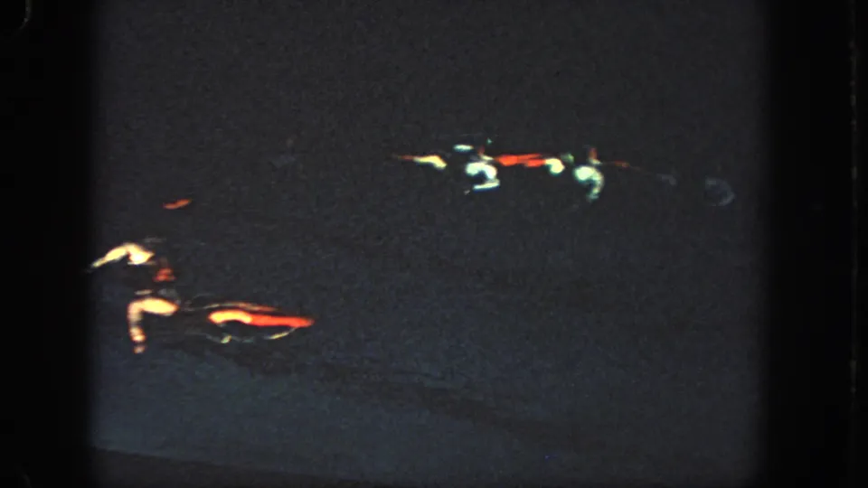 Near the end of the clip, two swimmers are partly illuminated in the dark pool, with red and white costume details standing out in the 8mm frame.