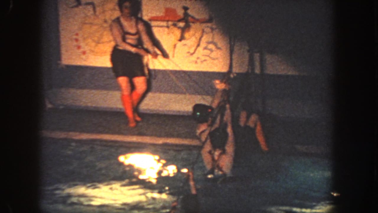 Grainy 1951 Georgia 8mm frame of indoor pool activity with a standing figure and swimmer near the edge.