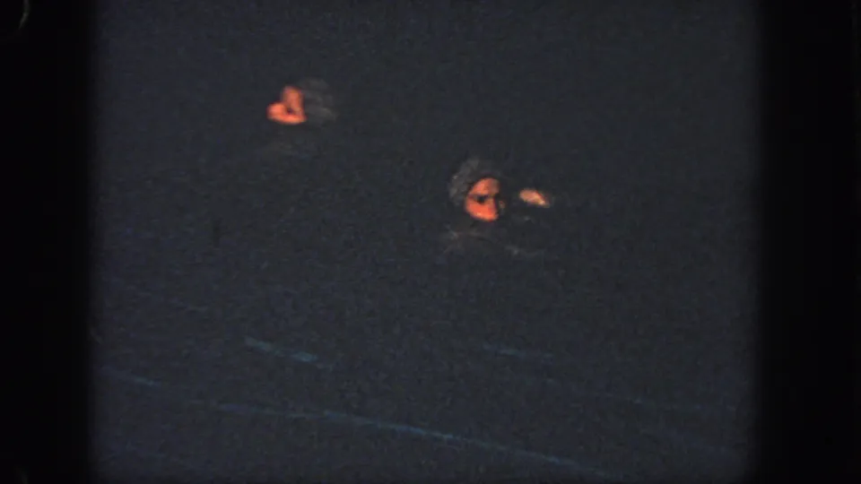 Two women in swim caps are visible in a dark pool, separated in the frame as part of a synchronized swimming movement.