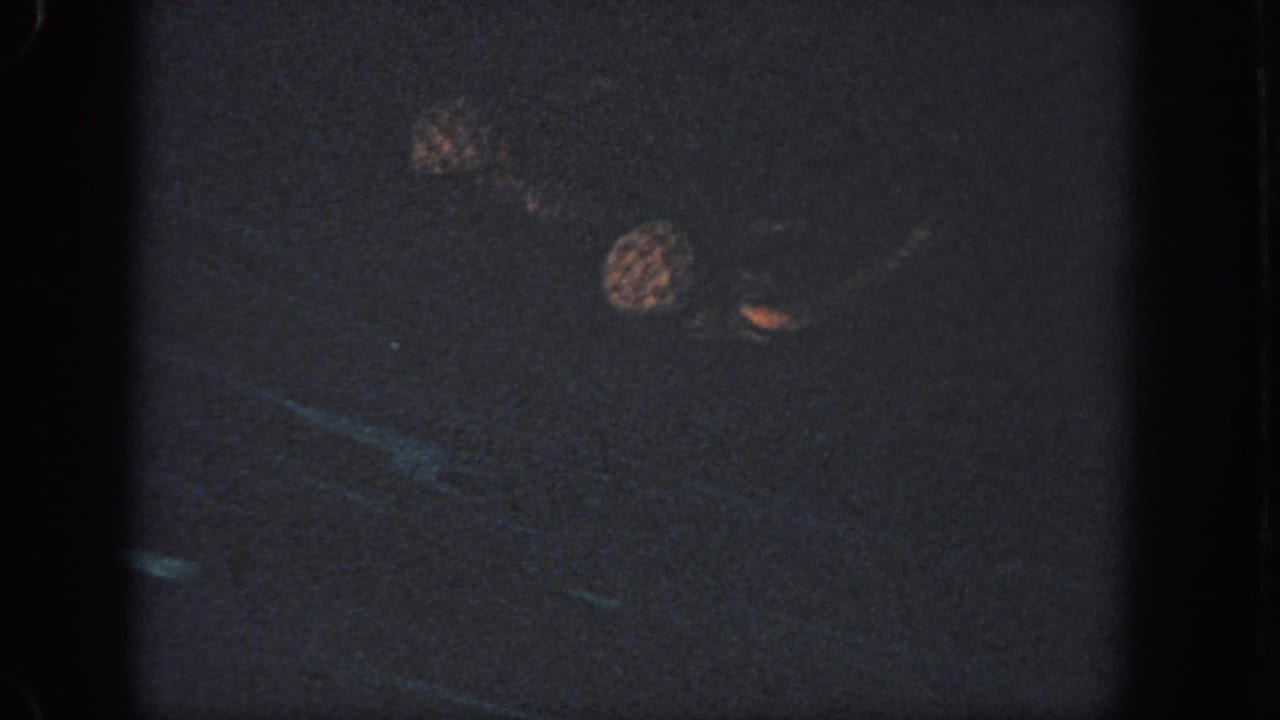Two women remain mostly submerged in the pool, with swim caps and a small raised hand visible in the dark 8mm frame.