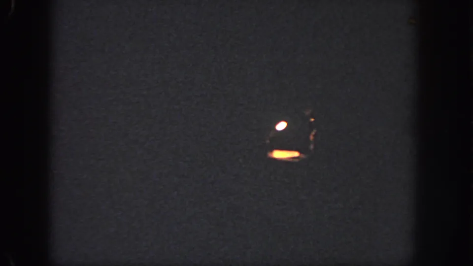 Low-light Georgia 8mm frame showing a small bright shape near dark water or ground, with surrounding grain and heavy shadow.
