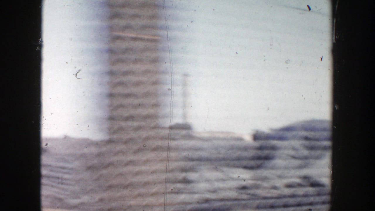 1956 Grand Rapids home-movie frame with motion-blurred construction materials, pale sky, and distant industrial forms from a passing vehicle.