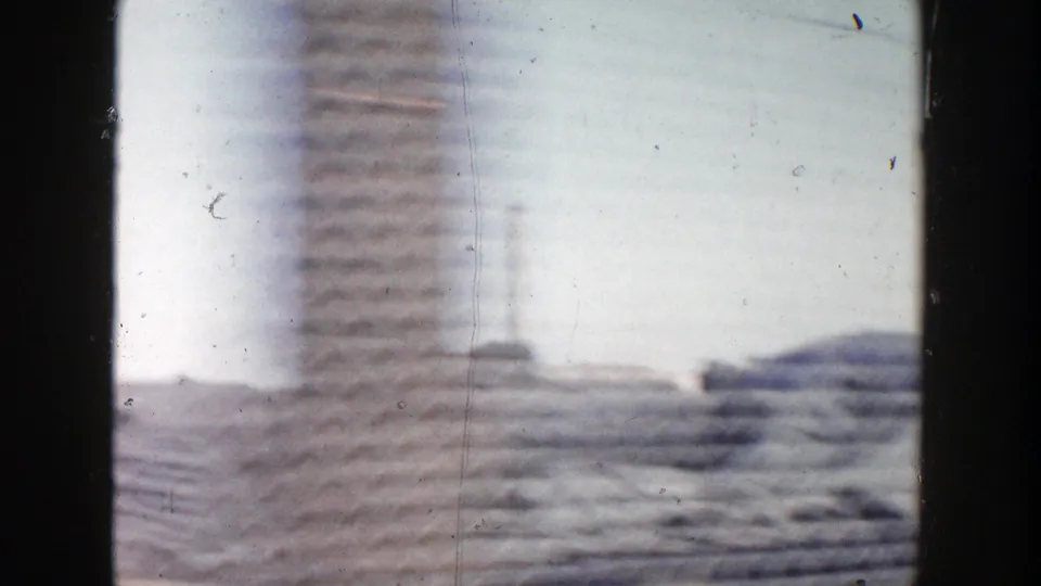 1956 Grand Rapids home-movie frame with motion-blurred construction materials, pale sky, and distant industrial forms from a passing vehicle.