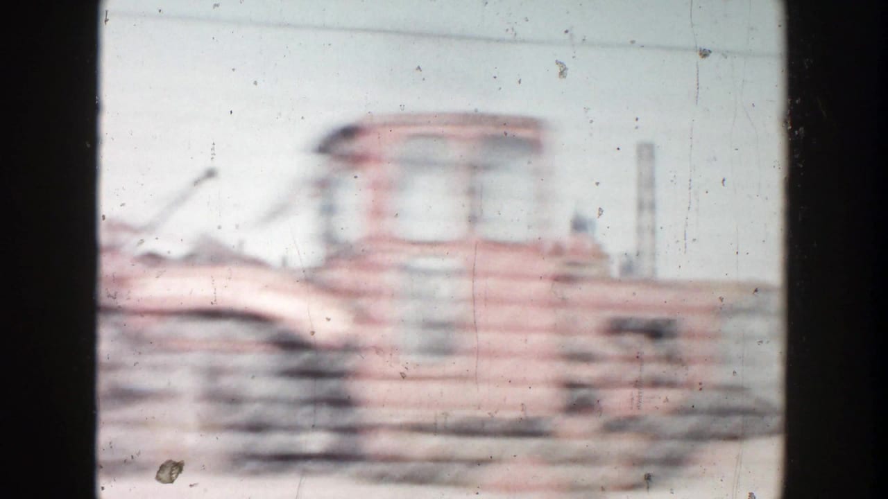 1956 Grand Rapids frame showing a motion-blurred reddish construction vehicle or equipment form near stacked materials under a pale sky.