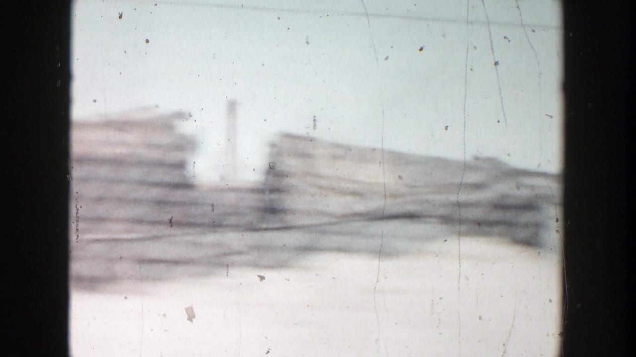 Final 8mm frame from the moving vehicle, showing hazy stacked materials, low industrial forms, and vertical pipes or smokestacks in Grand Rapids.