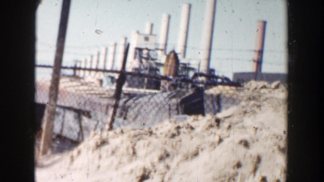 Dirt pile beside a chain-link fence with factory smokestacks in the distance.