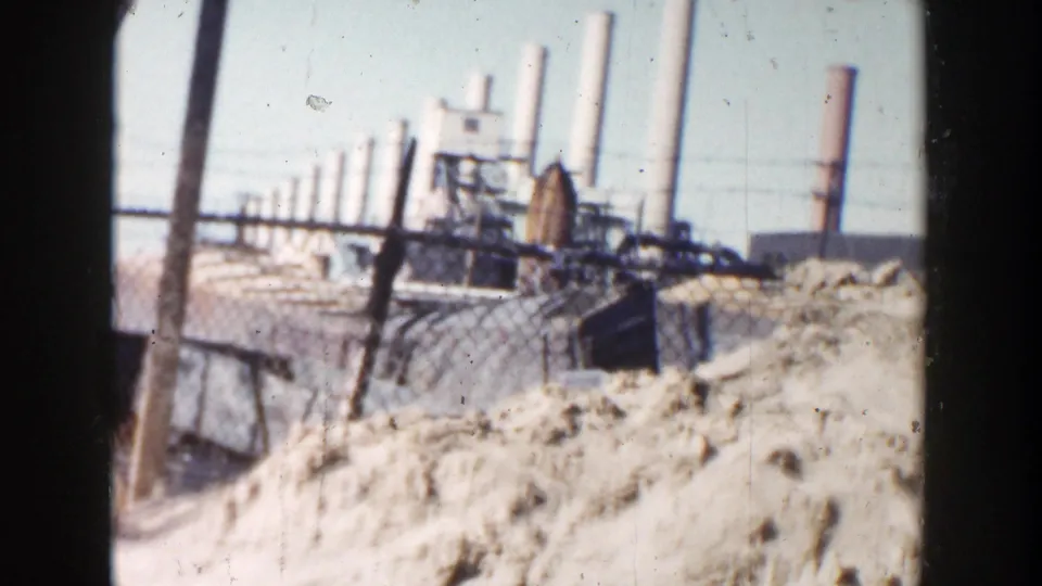 1956 Grand Rapids 8mm frame showing a dirt pile, chain-link fence, industrial structures, and distant smokestacks under bright sky.
