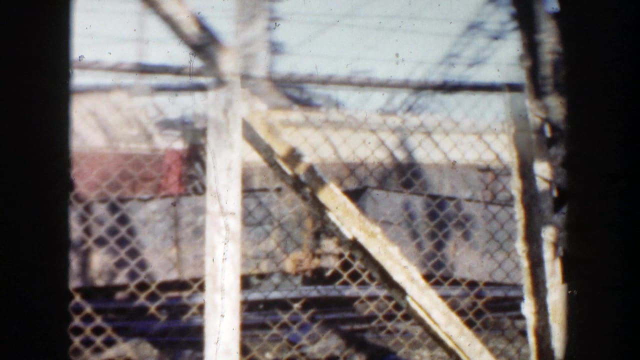 1956 Grand Rapids 8mm frame showing a chain-link gate, diagonal beam, dirt pile, and industrial background during the pan.