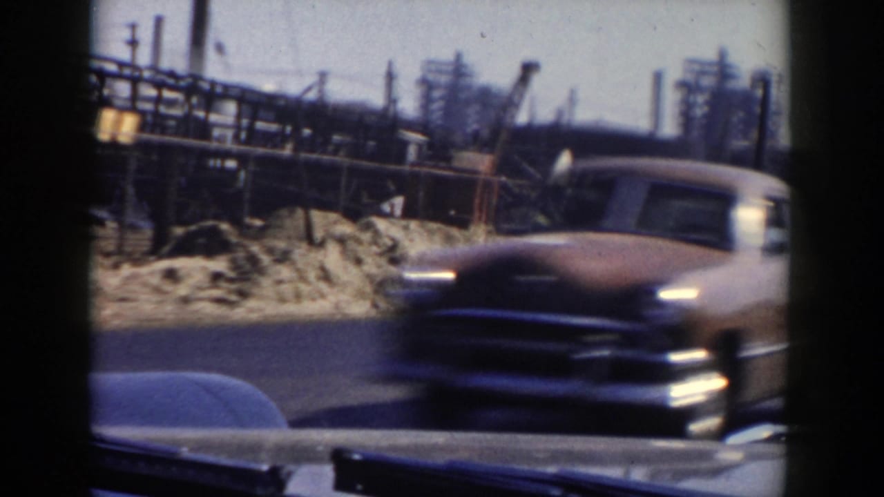 A rounded mid-century car passes a roadside industrial or construction area in Grand Rapids, Michigan, seen through a vehicle window.