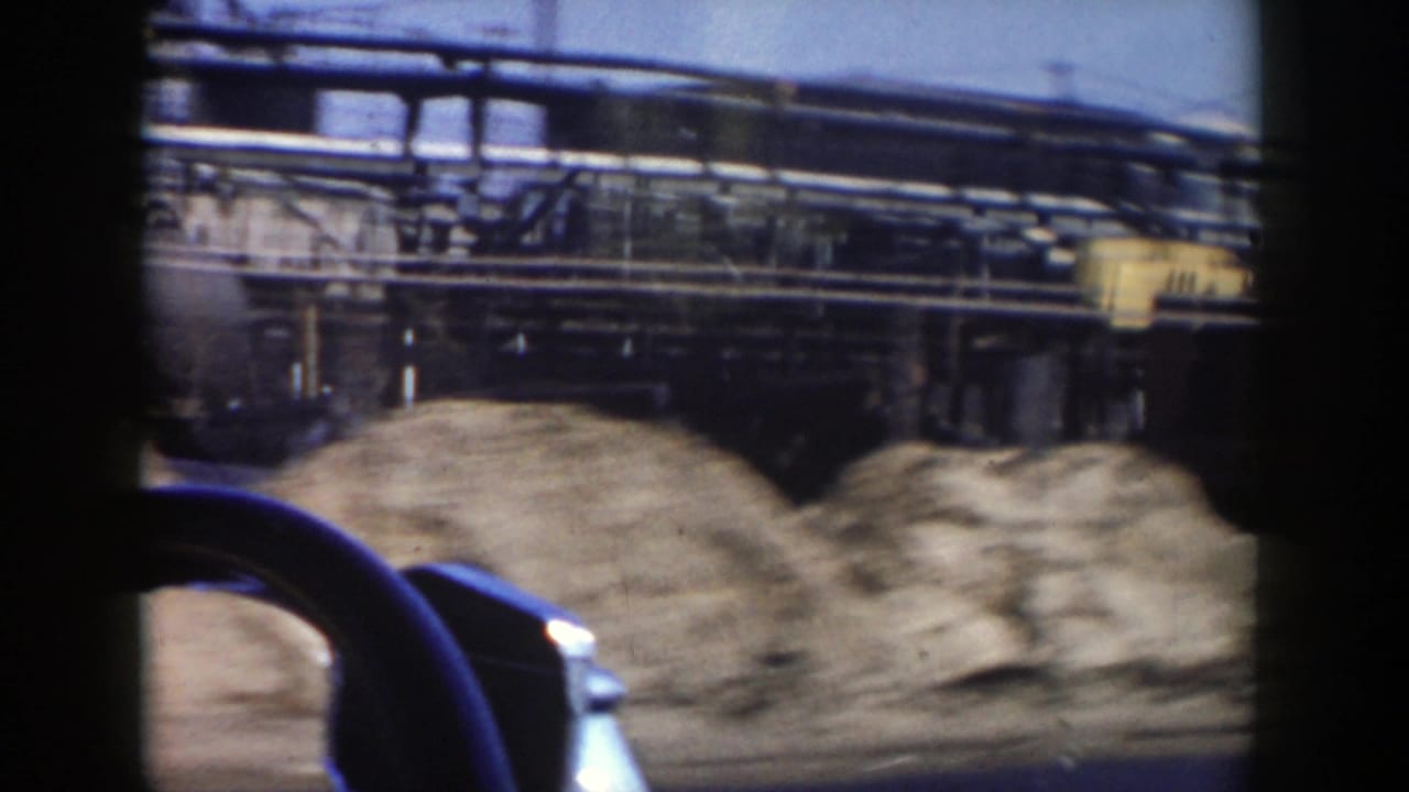 A 1956 Grand Rapids frame shows dirt piles and industrial structures beyond the road, with part of the filming vehicle visible at the bottom.