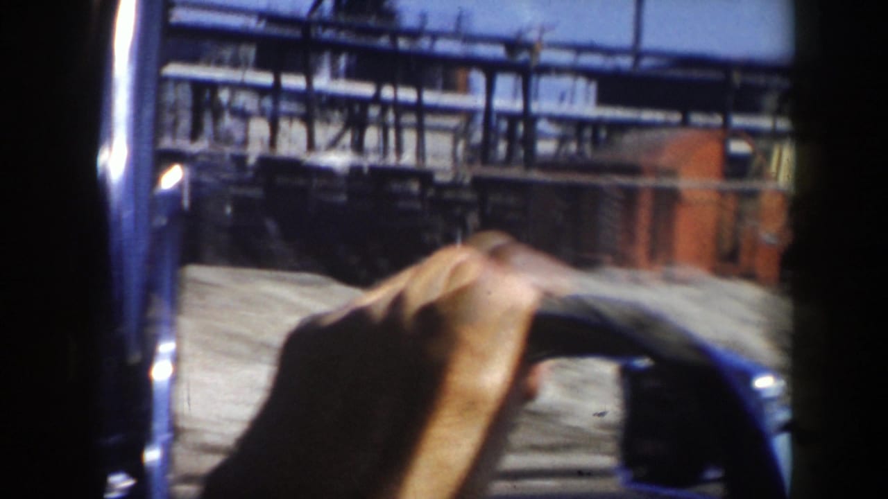 Hand on a steering wheel seen through a vehicle window near industrial structures.