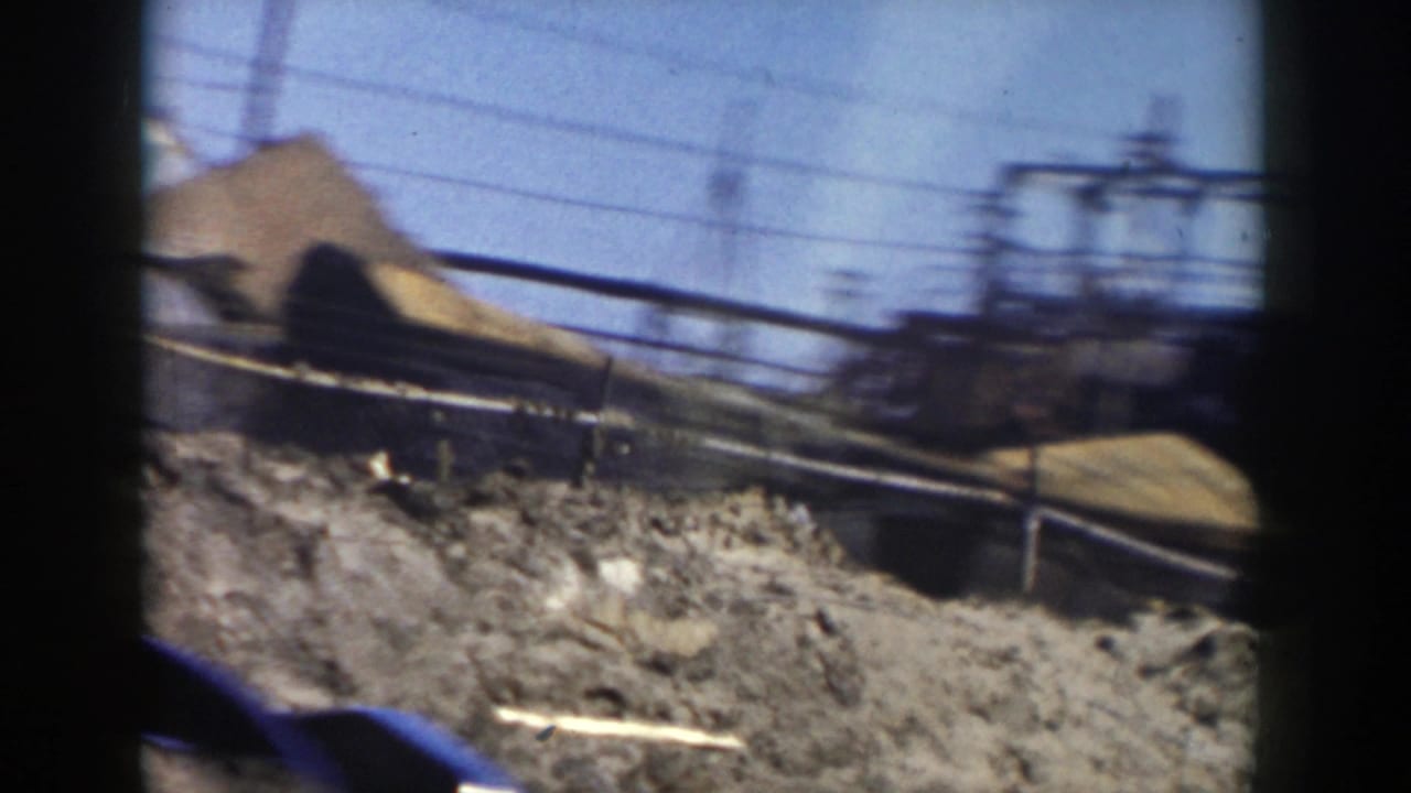 Passing 1956 Grand Rapids view with dirt piles, power lines, railings, and industrial framework under a bright sky.