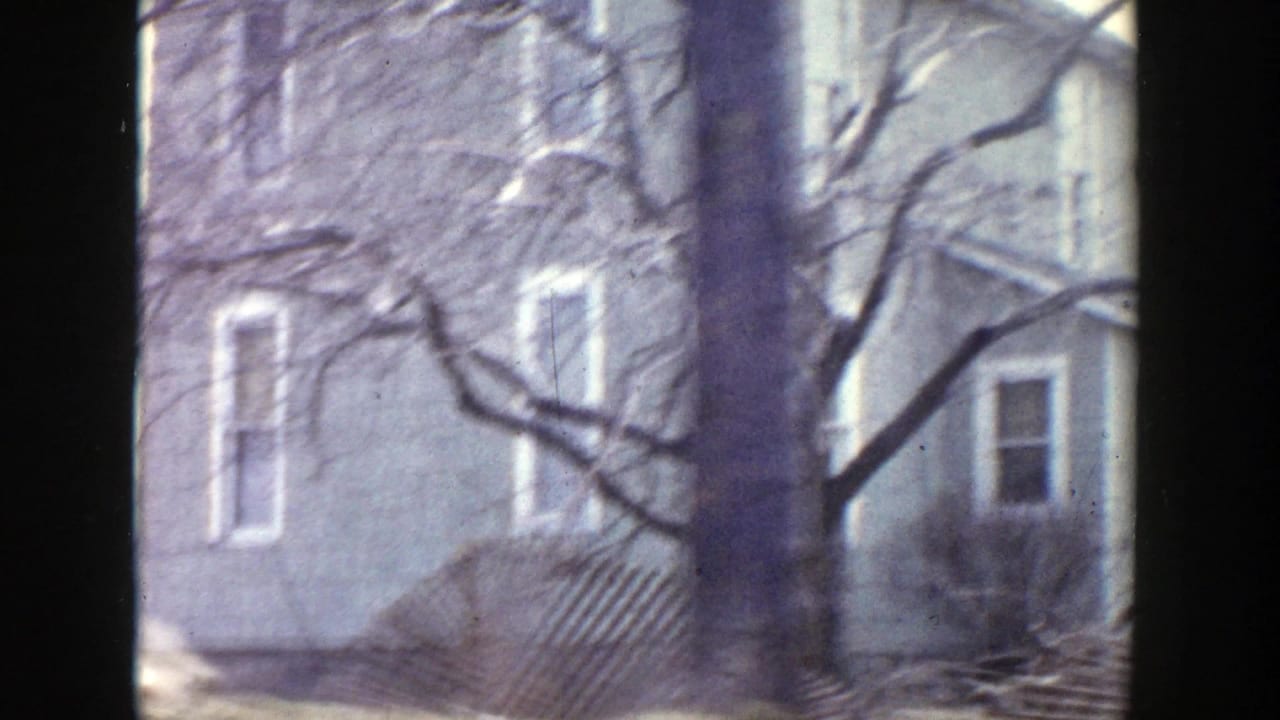 A Grand Rapids residential exterior appears through the moving 8mm camera, with bare trees, windows, and a low porch or stair rail.