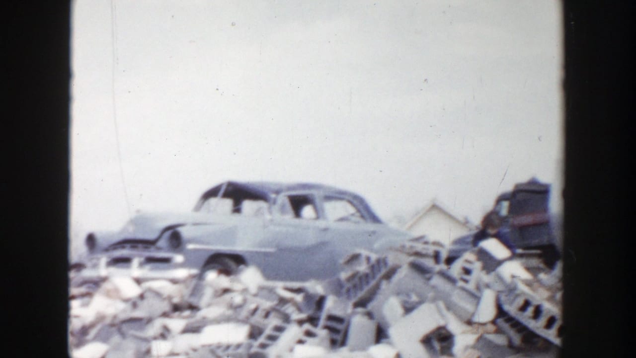 1956 Grand Rapids home-movie frame focused on a weathered sedan beside broken bricks, debris, and distant small buildings.