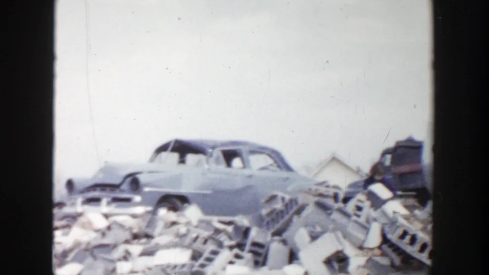 1956 Grand Rapids home-movie frame focused on a weathered sedan beside broken bricks, debris, and distant small buildings.