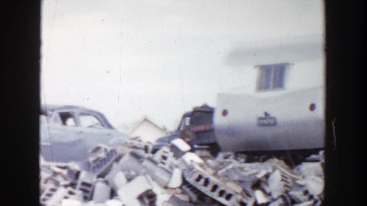 1956 Grand Rapids 8mm frame showing a small trailer, a partly visible sedan, and piles of discarded masonry in a junkyard setting.