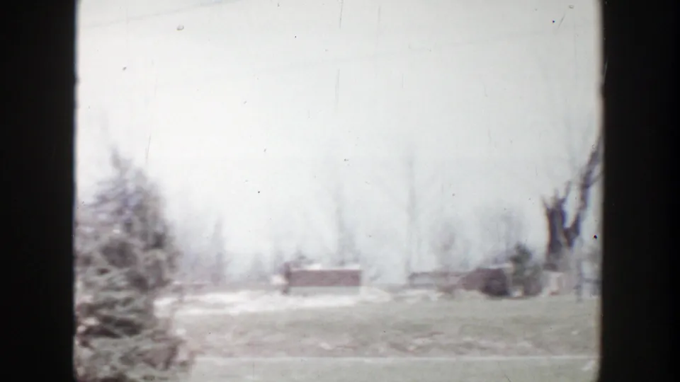 1956 Grand Rapids frame showing an open yard, leafless trees, distant structure, and remaining debris after visible damage.
