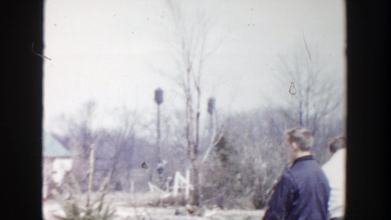 A man in a dark jacket stands near bare trees and snowy ground.