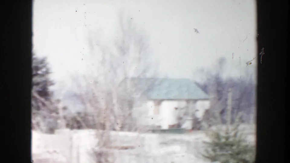 A small light-colored house and leafless trees appear beyond snow-dusted ground in the 1956 Grand Rapids home movie.