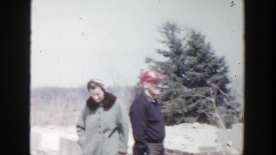 A woman in a light winter coat and a man in a red cap stand outdoors in Grand Rapids winter footage from 1956.