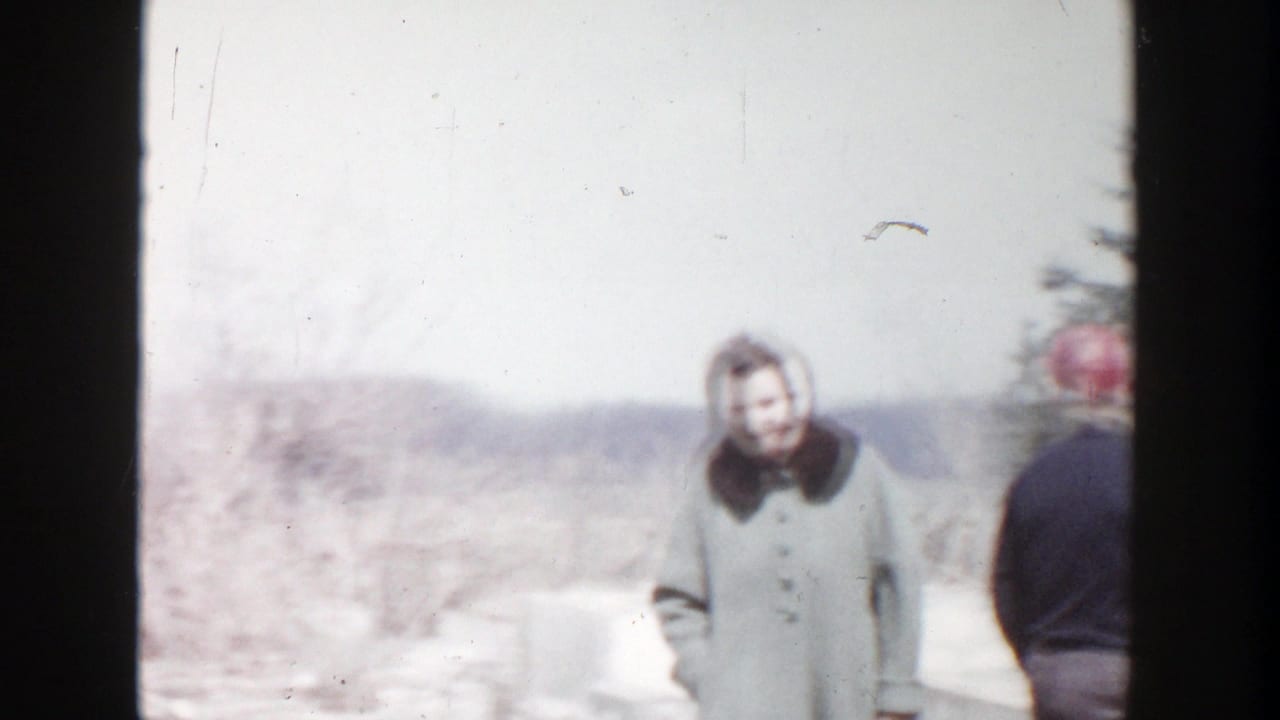 In 1956 Grand Rapids home-movie footage, a woman in a winter coat and a man in a red cap stand near snowy residential fire debris.