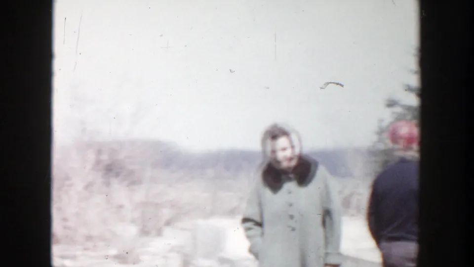In 1956 Grand Rapids home-movie footage, a woman in a winter coat and a man in a red cap stand near snowy residential fire debris.