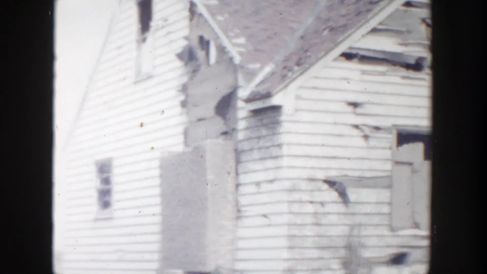 A side view of the damaged Grand Rapids house shows charred exterior wall sections, broken siding, and roof damage.