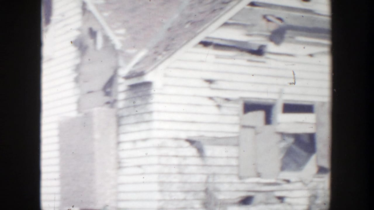 1956 Grand Rapids home-movie frame showing a damaged white clapboard house with broken upper windows, torn siding, and roof damage.