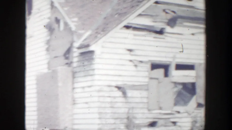 1956 Grand Rapids home-movie frame showing a damaged white clapboard house with broken upper windows, torn siding, and roof damage.
