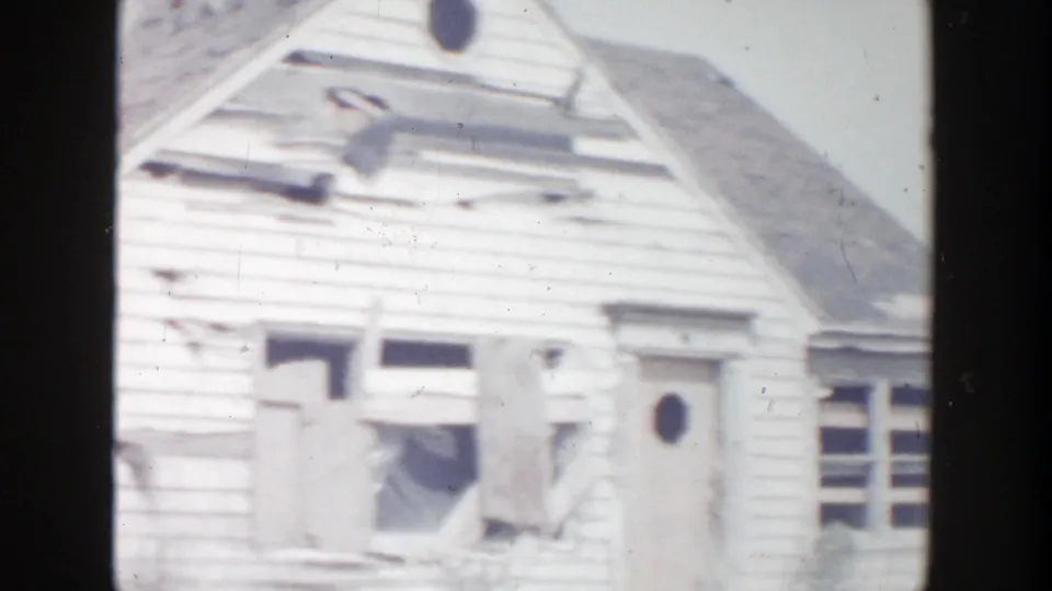 1956 Grand Rapids frame showing the damaged facade of a white clapboard house, including broken windows, torn siding, and a door with a round window.