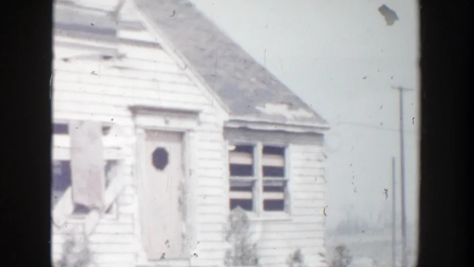 1956 Grand Rapids home-movie frame showing the front corner of a damaged white house with a door, window, and torn material near the entrance.
