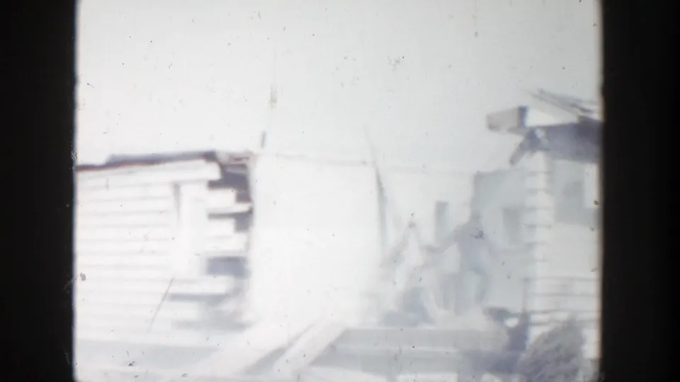 1956 Grand Rapids frame showing the side of a damaged white house, with missing siding, exposed structure, debris, and bright overexposed sky.