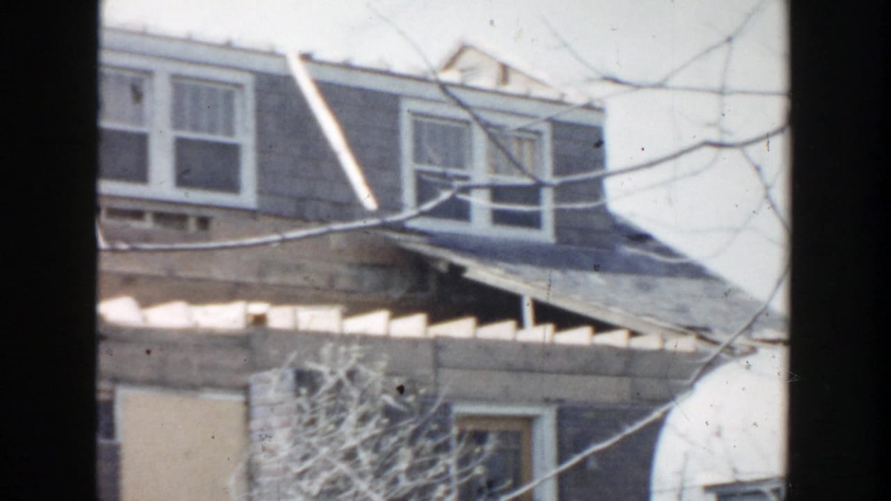 1956 Grand Rapids home-movie frame showing a weathered house exterior, dormer windows, roofline, and bare tree branches.