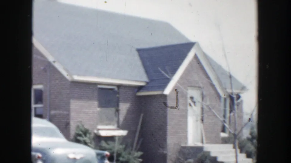1956 Grand Rapids frame showing a brick residential house with a gray roof, front steps, shrubs, and part of a parked sedan.