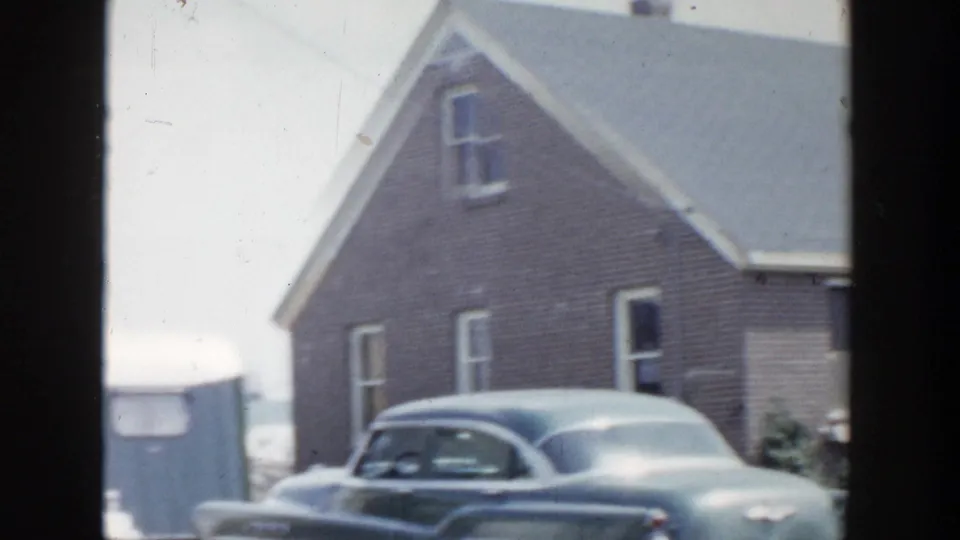 1956 Grand Rapids home-movie view of a brick house side wall, gable roof, windows, and a rounded sedan parked nearby.