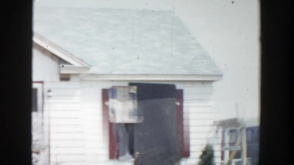 1956 Grand Rapids home-movie frame showing a white house with red trim, a dark damaged opening, and soft 8mm exposure.