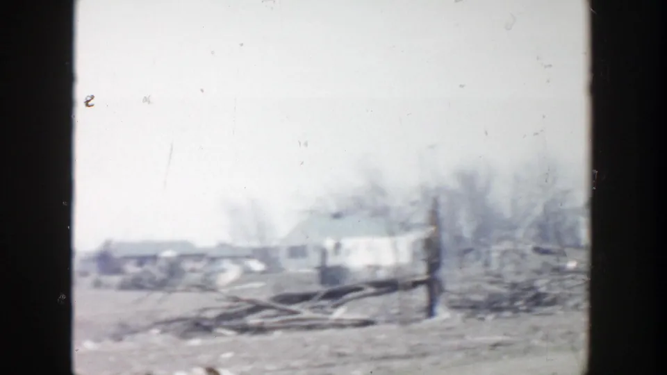 1956 Grand Rapids frame showing a wider residential aftermath view with debris, fence posts, bare trees, and small distant houses.