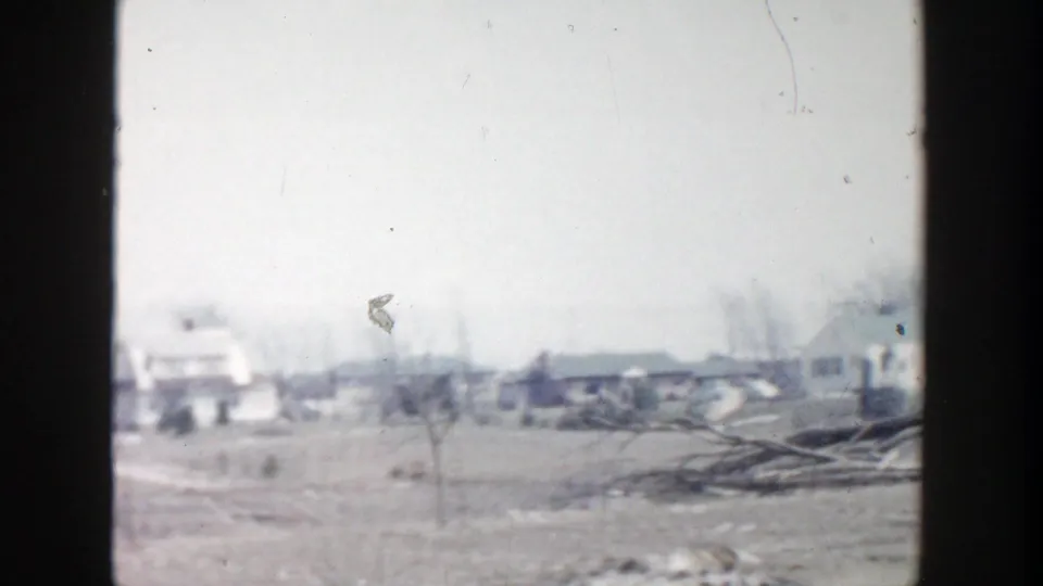 1956 Grand Rapids-area home-movie frame showing barren farm ground, distant low buildings, bare trees, and debris under an overcast sky.