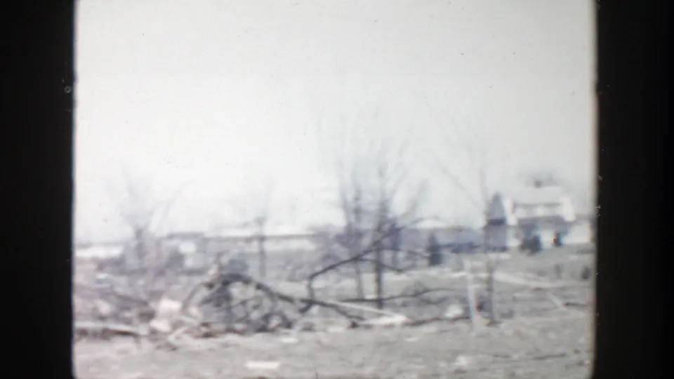 1956 Grand Rapids-area frame with fallen branches, bare trees, open farm ground, and a distant low structure in soft 8mm focus.