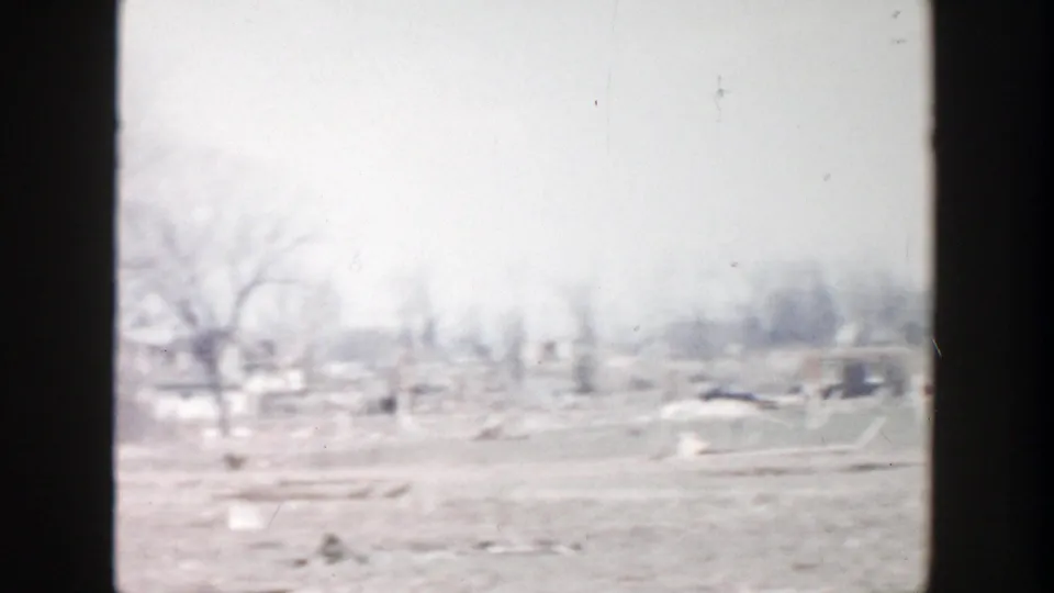 1956 Grand Rapids-area frame of an open rural field with bare trees, scattered debris, and distant low structures beneath a gray sky.